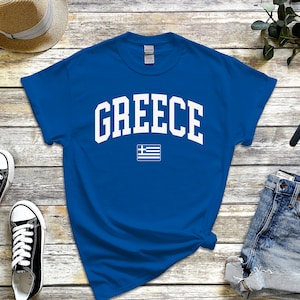Może przedstawiać: Koszulka w kolorze królewskiej niebieskości z białym napisem "GREECE" w łukowatym kroju czcionki. Pod napisem znajduje się mała biała grecka flaga.