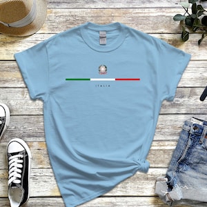 Puede incluir: Camiseta azul claro con una franja verde, blanca y roja a trav&eacute;s del pecho. La franja presenta el escudo de armas italiano y la palabra "ITALIA" debajo.