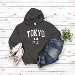 May include: A charcoal gray hooded sweatshirt with the word "TOKYO" in white letters and a Japanese flag with the text "日本" underneath. The sweatshirt is laid flat on a white wood background with a pair of white sneakers and blue jeans.