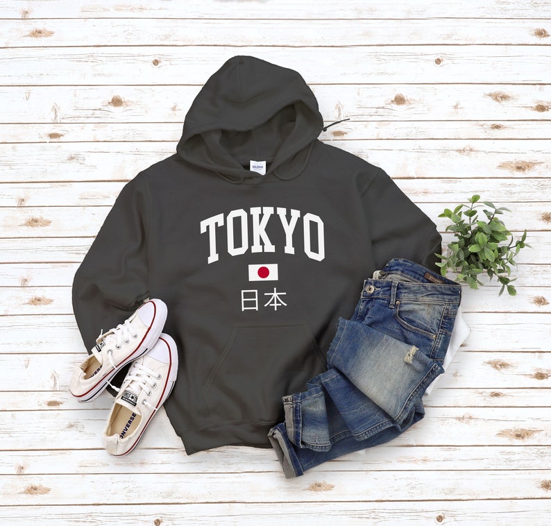 May include: A charcoal gray hooded sweatshirt with the word "TOKYO" in white letters and a Japanese flag with the text "日本" underneath. The sweatshirt is laid flat on a white wood background with a pair of white sneakers and blue jeans.
