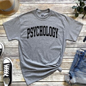 Puede incluir: Una camiseta gris con la palabra "PSYCHOLOGY" impresa en letras negras en una fuente arqueada.