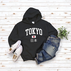 May include: Black hooded sweatshirt with white text that says "TOKYO" and a Japanese flag with the text "日本" underneath. The sweatshirt is laid out on a white wood background with a pair of white sneakers and blue jeans.