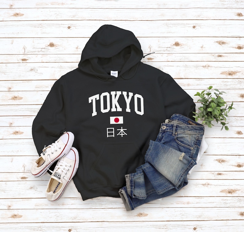 May include: Black hooded sweatshirt with white text that says "TOKYO" and a Japanese flag with the text "日本" underneath. The sweatshirt is laid out on a white wood background with a pair of white sneakers and blue jeans.