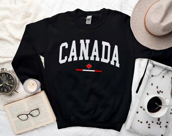 Pode incluir: Camisola de moletom preta com texto branco que diz "CANADA" e um gráfico de folha de bordo vermelha.