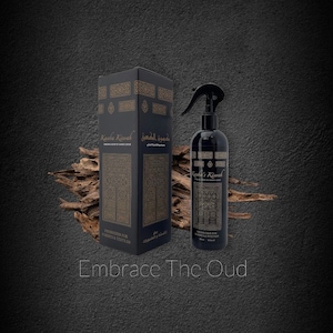 May include: A black spray bottle with a gold label that reads "Kaaba Kiswah" and "Freshener for Fabrics & Textiles". The bottle is sitting on a wooden surface with a black box behind it. The box also has a gold label that reads "Kaaba Kiswah" and "Freshener for Fabrics & Textiles". The text "Embrace The Oud" is written in white letters on a black background.