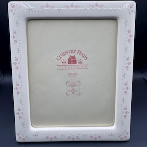 May include: A white ceramic picture frame featuring a border decorated with small pink hearts and green leaf accents. The centre insert displays the text Country Haus Collection, Ceramic 8 x 10, and Another Quality Crafted Frame AK International.