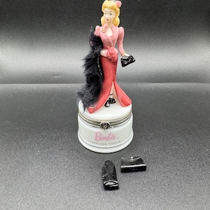 May include: A porcelain figurine of a woman in a pink dress and black fur stole, holding a black purse. The figurine is on a white base with the word "Barbie" and "Fabulous Forties" printed on it. A pair of black gloves and a black purse are next to the base.