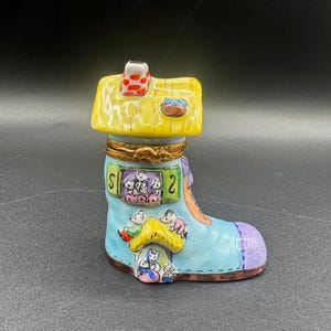 May include: A whimsical, hand-painted ceramic boot-shaped trinket box. The boot is light blue with a purple toe and features a yellow house-shaped lid with a red chimney. Adorable cartoon characters are painted on the boot.