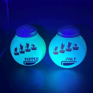 Puede incluir: Un par de saleros y pimenteros vintage. Los saleros son redondos con tapas azules y brillan en verde brillante. La palabra "PEPPER" está impresa en uno, y "SALT" en el otro. Cada salero tiene ilustraciones de plantas.