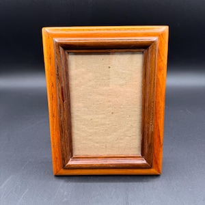 May include: A rectangular wooden picture frame featuring a two-tone design with a lighter orange-brown outer border and a darker reddish-brown inner border. The frame has a smooth, polished finish and contains a plain, light tan cardboard insert.