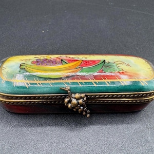 Könnte beinhalten: Eine rechteckige Schmuckschatulle mit Klappdeckel. Die Schatulle ist mit einem Fruchtdesign bemalt, das Bananen, Trauben und Wassermelonen zeigt. Die Schatulle hat einen grünen und goldenen Rand und einen bronzefarbenen Traubenverschluss.