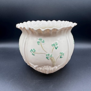May include: A cream-colored ceramic planter with a ruffled, scalloped rim and vertical fluted detailing. The exterior features a raised, hand-painted green shamrock pattern with delicate stems, creating a textured surface on the rounded body.