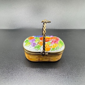 Könnte beinhalten: Eine ovale Schmuckschatulle mit Klappdeckel und dekorativem Griff. Der Deckel ist mit einem farbenfrohen Blumendesign mit roten, lila und orangen Blumen bemalt. Die Schatulle hat ein Korbgeflechtmuster an den Seiten und einen goldfarbenen Metallgriff.