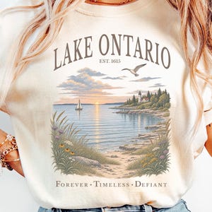 May include: An ivory short-sleeved t-shirt featuring a graphic print of Lake Ontario. The design includes the text "LAKE ONTARIO EST. 1615" above a landscape illustration of a lake, sailboat, lighthouse, and seagull, with "FOREVER • TIMELESS • DEFIANT" below.