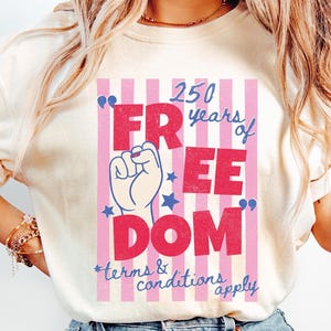 May include: Ivory-colored t-shirt with a graphic that reads "250 years of FREEDOM" in red and blue, with a raised fist illustration. The design is set against a pink and white striped background. The text "terms & conditions apply" is at the bottom.