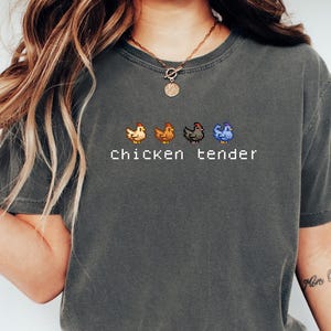 May include: Dark grey t-shirt featuring the text "Chicken tender" and four pixelated chicken illustrations in orange, brown, black, and blue. The shirt has a crew neck and short sleeves.