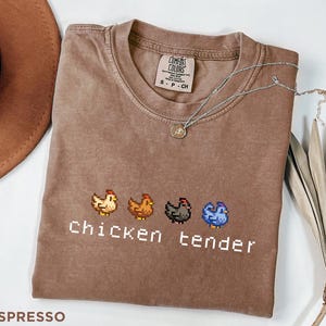 May include: Espresso-coloured t-shirt featuring a pixelated chicken design and the words "chicken tender" in white. The shirt has a crew neck and short sleeves. A silver necklace with a small pendant rests on the shirt.