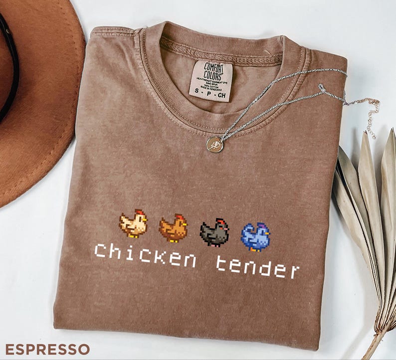 May include: Espresso-coloured t-shirt featuring a pixelated chicken design and the words "chicken tender" in white. The shirt has a crew neck and short sleeves. A silver necklace with a small pendant rests on the shirt.