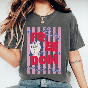 May include: Dark gray t-shirt with a graphic that reads "250 years of FREEDOM" in red letters. The design includes a raised fist, stars, and pink and white stripes. The text "*terms & conditions apply" is in blue.
