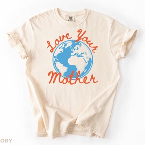 May include: An ivory t-shirt featuring the words "Love Your Mother" in red script, with a blue globe graphic. The shirt has a crew neck and rolled-up sleeves. The colour is ivory.