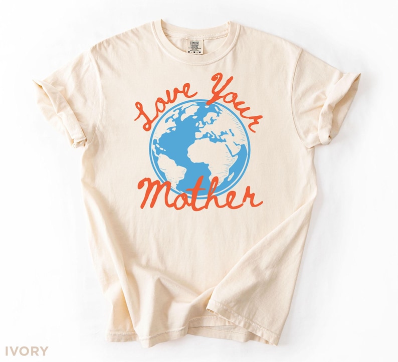 May include: An ivory t-shirt featuring the words "Love Your Mother" in red script, with a blue globe graphic. The shirt has a crew neck and rolled-up sleeves. The colour is ivory.