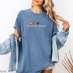 May include: Blue jean coloured t-shirt with the text "chicken tender" and pixelated chicken graphics in orange, brown, black, and blue. The shirt has rolled-up sleeves and is worn with a light blue denim jacket.