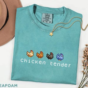 May include: Seafoam green t-shirt featuring a pixelated chicken design and the text "Chicken tender". A gold star pendant necklace is on the shirt. The shirt is a size XL-TG-EG.