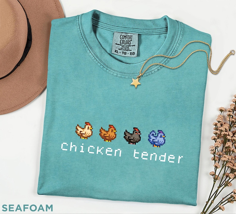 May include: Seafoam green t-shirt featuring a pixelated chicken design and the text "Chicken tender". A gold star pendant necklace is on the shirt. The shirt is a size XL-TG-EG.