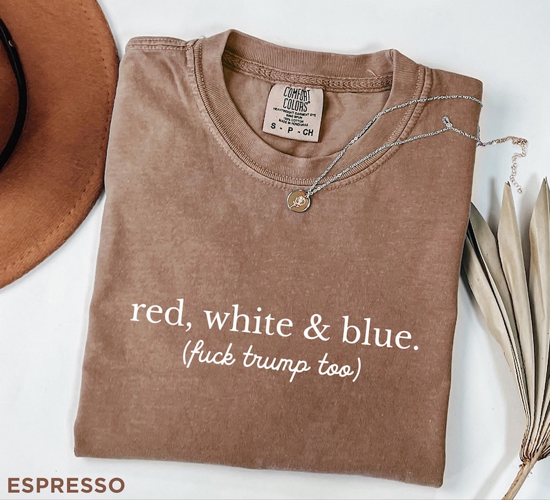 May include: Espresso-colored t-shirt with the text "red, white & blue. (fuck trump too)". The shirt has a crew neck and a small tag on the upper chest. A silver necklace with a pendant is draped across the shirt.