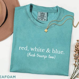 May include: Seafoam green t-shirt with the text "red, white & blue. (fuck trump too)". A gold star pendant necklace rests on the shirt. The shirt has a Comfort Colors tag and is size XL-TG-EG.