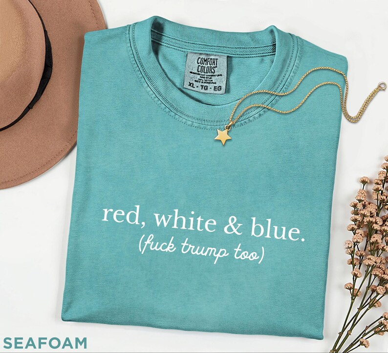 May include: Seafoam green t-shirt with the text "red, white & blue. (fuck trump too)". A gold star pendant necklace rests on the shirt. The shirt has a Comfort Colors tag and is size XL-TG-EG.
