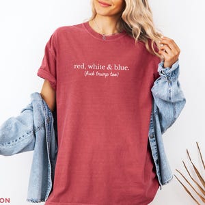 May include: Crimson-colored t-shirt with the text "red, white & blue. (fuck trump too)". The shirt has short sleeves and a relaxed fit. A light blue denim jacket is draped over the shoulders.