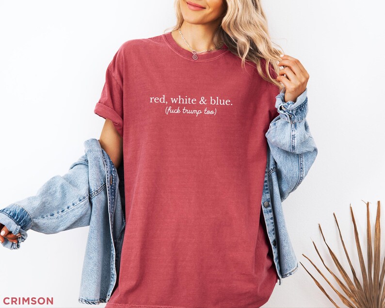 May include: Crimson-colored t-shirt with the text "red, white & blue. (fuck trump too)". The shirt has short sleeves and a relaxed fit. A light blue denim jacket is draped over the shoulders.