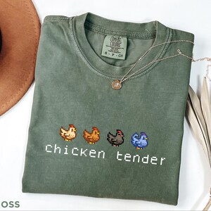 May include: A moss green t-shirt featuring pixelated chicken designs and the words "Chicken tender" in white. A silver necklace with a pendant rests on the shirt. The t-shirt is folded on a white surface.