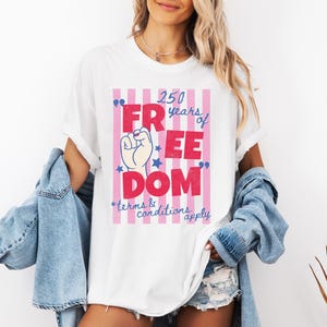 May include: White t-shirt with a graphic that reads "250 years of FREEDOM" in red and blue, with a raised fist and stars. The design is set against a pink and white striped background. The text "terms & conditions apply" is also included.