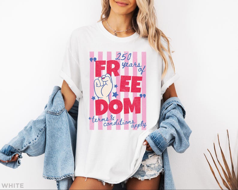May include: White t-shirt with a graphic that reads "250 years of FREEDOM" in red and blue, with a raised fist and stars. The design is set against a pink and white striped background. The text "terms & conditions apply" is also included.