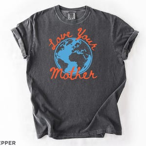 May include: A dark grey t-shirt with rolled sleeves. The shirt displays a blue and orange graphic of the Earth with the words "Love Your Mother" in orange script. The shirt is made of a soft, comfortable material.