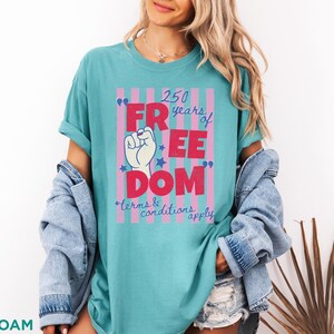May include: Seafoam green t-shirt with a graphic that reads "250 years of FREEDOM" in red letters. The design includes a raised fist, stars, and the text "terms & conditions apply". The shirt is a casual style.