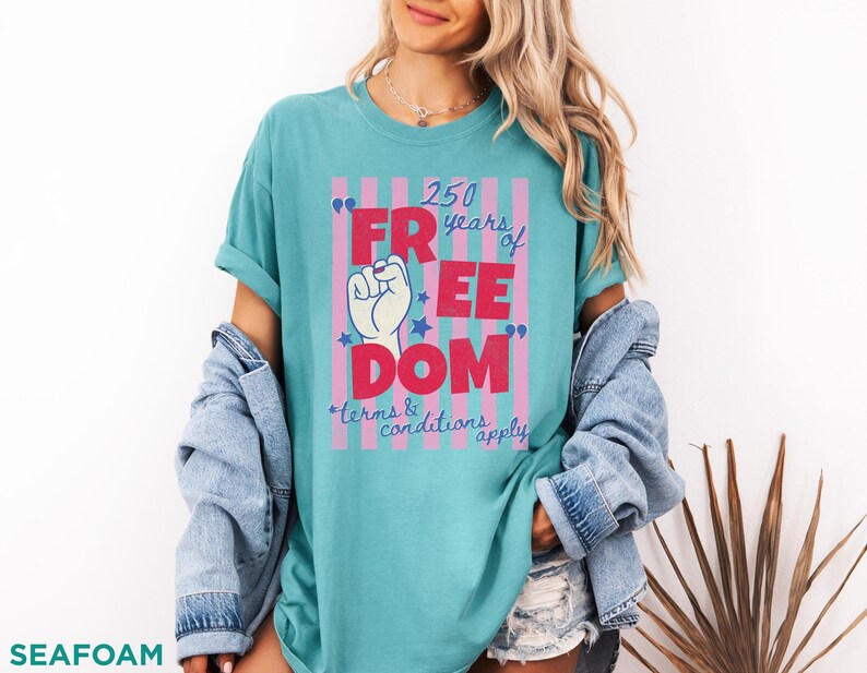 May include: Seafoam green t-shirt with a graphic that reads "250 years of FREEDOM" in red letters. The design includes a raised fist, stars, and the text "terms & conditions apply". The shirt is a casual style.