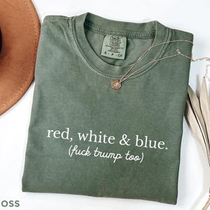 May include: A moss green t-shirt with the text "red, white & blue. (fuck trump too)". The shirt has a crew neck and short sleeves. A gold necklace with a pendant is draped on the shirt.