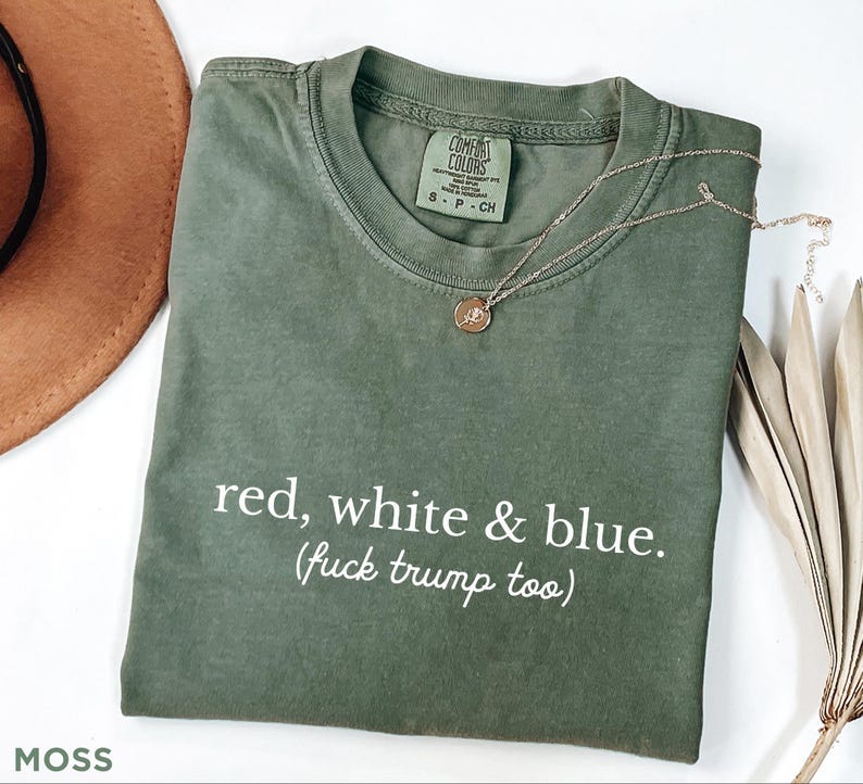 May include: A moss green t-shirt with the text "red, white & blue. (fuck trump too)". The shirt has a crew neck and short sleeves. A gold necklace with a pendant is draped on the shirt.