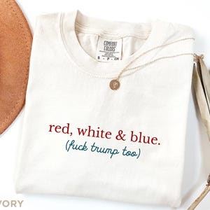 May include: Ivory-colored t-shirt with the text "red, white & blue. (fuck trump too)" in red and teal lettering. A gold necklace with a pendant is draped across the shirt. The shirt has a Comfort Colors tag.