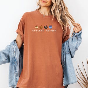 May include: A terracotta-coloured t-shirt featuring the words "chicken tender" and four pixelated chicken designs. The shirt is worn with a light blue denim jacket.