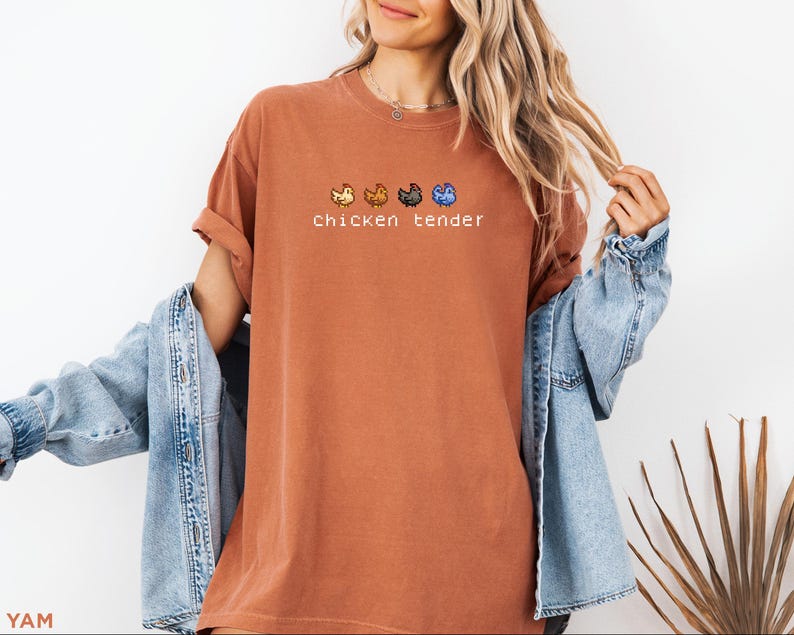 May include: A terracotta-coloured t-shirt featuring the words "chicken tender" and four pixelated chicken designs. The shirt is worn with a light blue denim jacket.