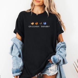 May include: Black t-shirt featuring the text "chicken tender" in white, with four pixelated chicken illustrations above. The chickens are in orange, brown, grey, and blue. The shirt is worn by a person with blonde hair and a denim jacket.