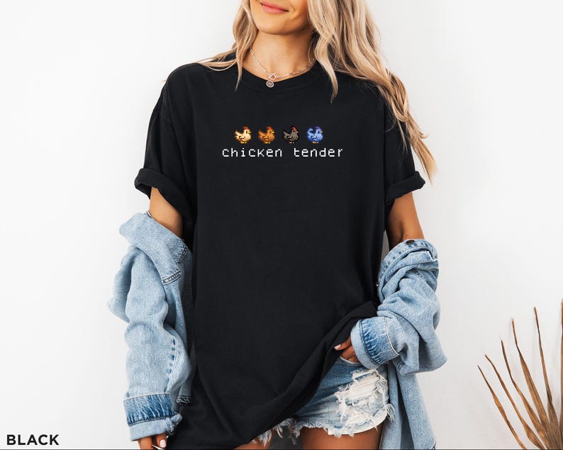 May include: Black t-shirt featuring the text "chicken tender" in white, with four pixelated chicken illustrations above. The chickens are in orange, brown, grey, and blue. The shirt is worn by a person with blonde hair and a denim jacket.