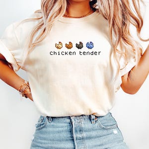 May include: Ivory t-shirt featuring the text "chicken tender" in a pixelated font. Above the text are four pixelated chickens in yellow, brown, grey, and blue. The shirt is worn with blue denim shorts.