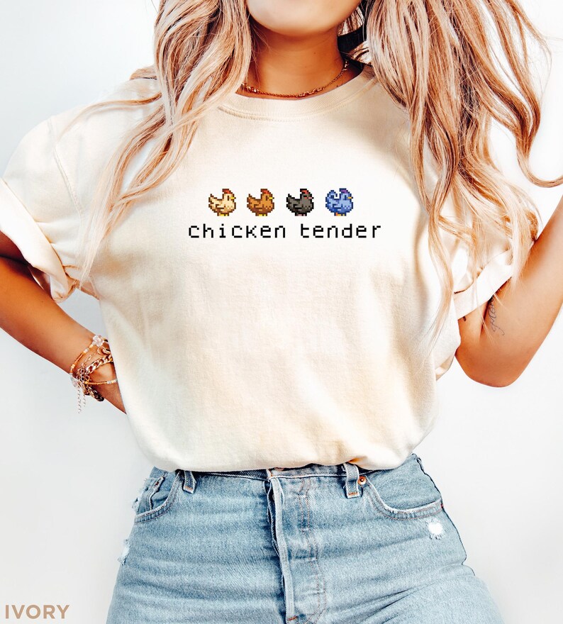 May include: Ivory t-shirt featuring the text "chicken tender" in a pixelated font. Above the text are four pixelated chickens in yellow, brown, grey, and blue. The shirt is worn with blue denim shorts.