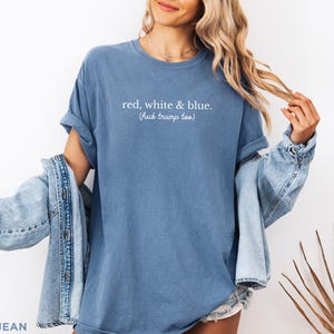 May include: A blue t-shirt with the text "red, white & blue. (fuck trump too)". The shirt is paired with a light blue denim jacket. The model is wearing a necklace and shorts.