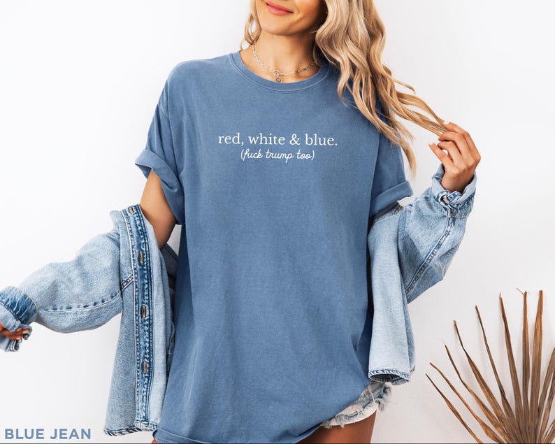 May include: A blue t-shirt with the text "red, white & blue. (fuck trump too)". The shirt is paired with a light blue denim jacket. The model is wearing a necklace and shorts.